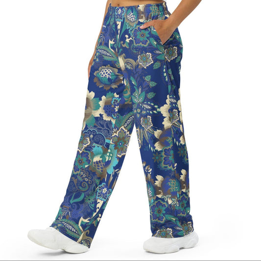 A woman wearing the rio flow joggers, wide-leg cut with a detailed botanical pattern in blue, green, and gold, paired with a white t-shirt and white sneakers, set against a plain white background.