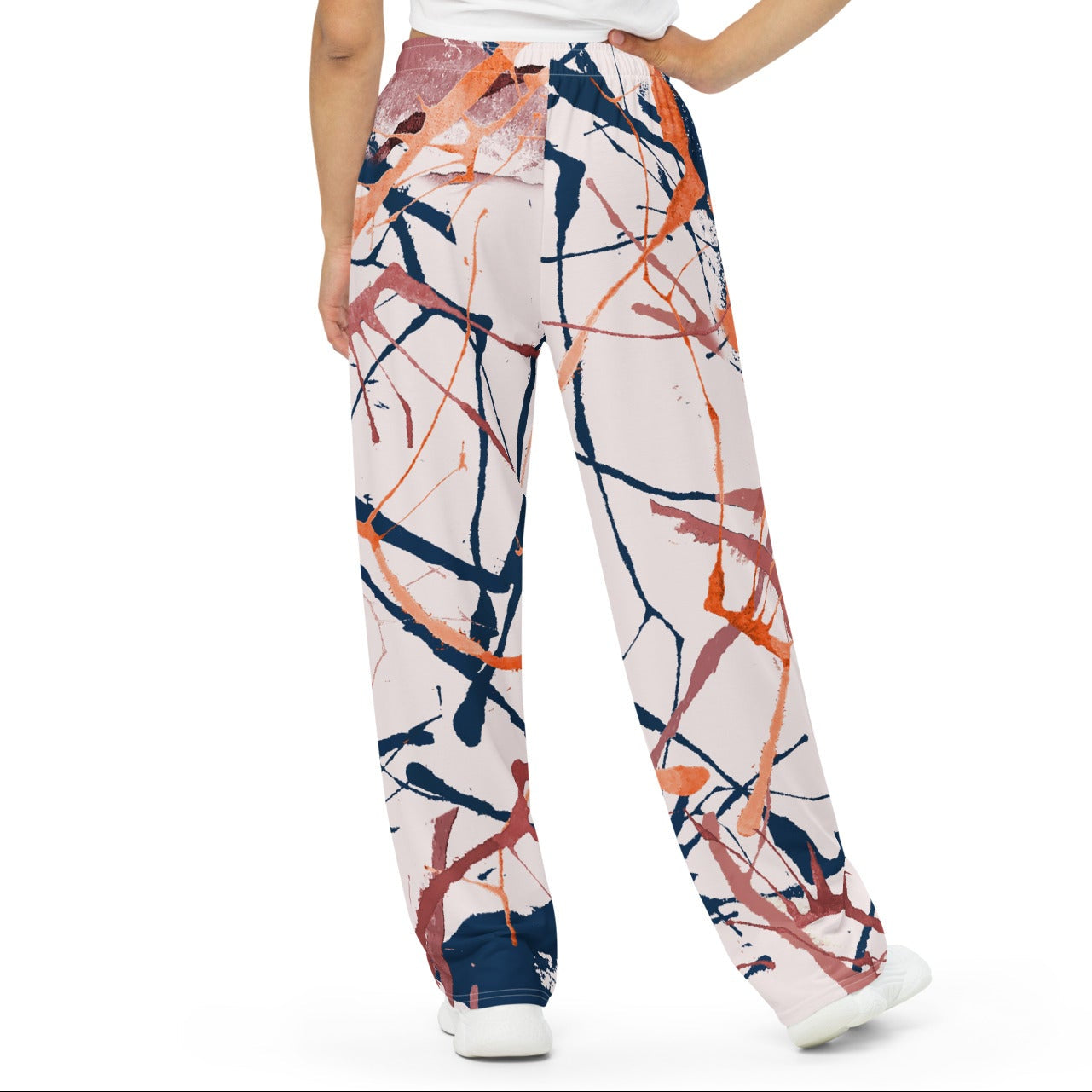 A woman is shown from the back wearing wide-leg trousers with an abstract paint splatter pattern in orange, navy, and pink on a white base, paired with a white t-shirt and white sneakers, against a plain white background.