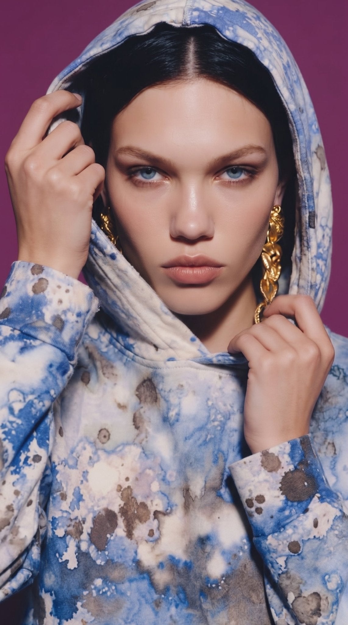 A close-up portrait of a woman wearing a hood over her head, featuring the same blue and white tie-dye hoodie. Chunky gold chain earrings dangle on either side of her face, and her hands lightly hold the edge of the hood near her cheeks against a rich purple background.