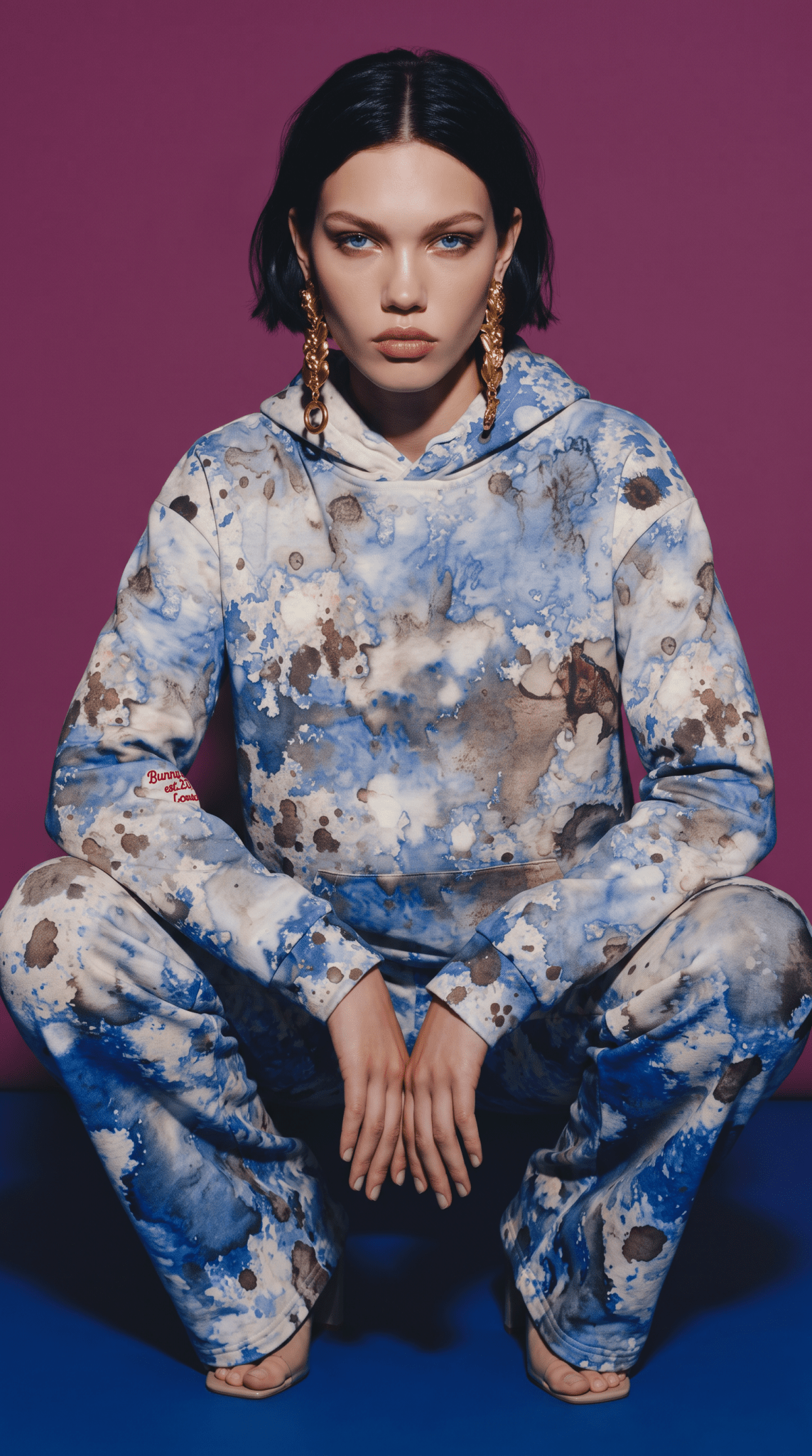 A model wearing the Amalfi Resort Hoodie and matching flow joggers in a matching blue and white abstract print hoodie and sweatpants set, posing in a crouched position against a solid purple background.