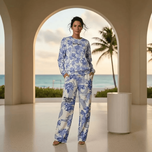 A woman stands in a modern, arched terrace overlooking a tropical beach at sunset, wearing the Saint Germain Glam Co-ord set. The outfit features a white jumper and matching wide-leg joggers adorned with an intricate, vintage-inspired blue floral and toile-like pattern and the text goal digger at the front.