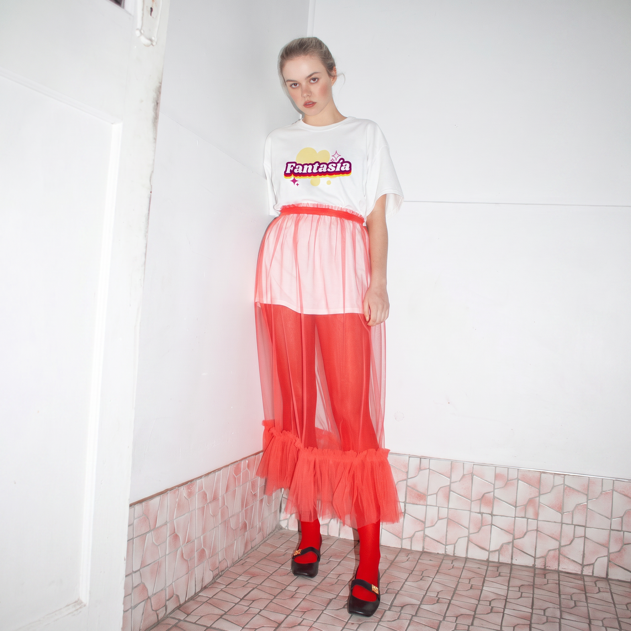 A model stands in a tiled room corner wearing the fantasia tee  a white graphic t-shirt with 'Fantasia' printed on the chest, a sheer red mesh skirt over red leggings and black shoes.