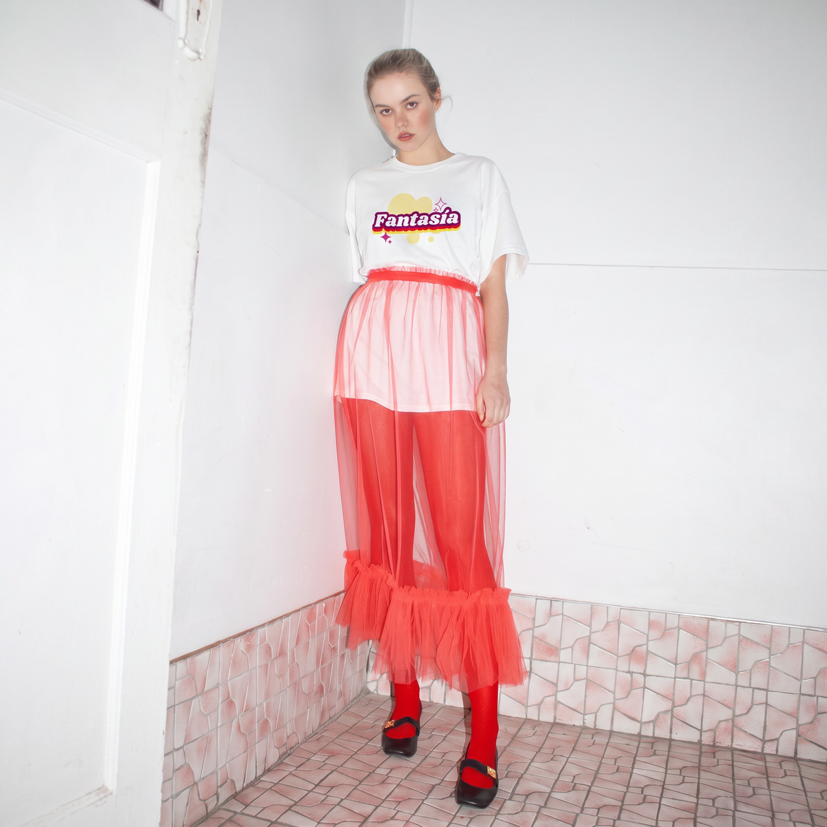 A model stands in a tiled room corner wearing the fantasia tee  a white graphic t-shirt with 'Fantasia' printed on the chest, a sheer red mesh skirt over red leggings and black shoes.