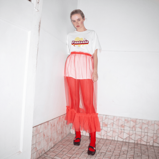 A model stands in a tiled room corner wearing the fantasia tee  a white graphic t-shirt with 'Fantasia' printed on the chest, a sheer red mesh skirt over red leggings and black shoes.
