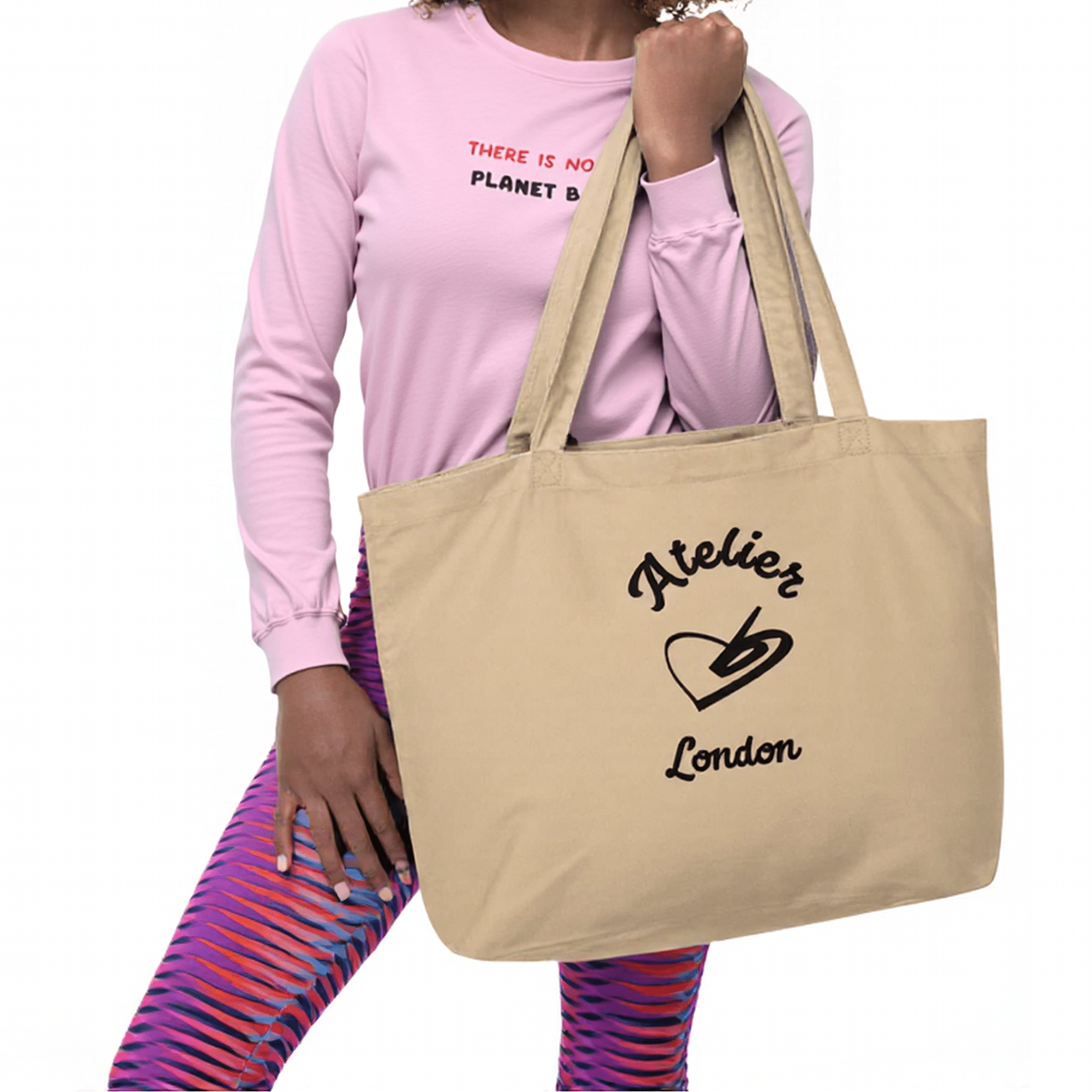 A model holding The Never Full Atelier Tote (featuring the Atelier Heart emblem) while wearing The Moorgate Sculpt Leggings (in the dynamic graphic arrow print) and The Manifesto Sweatshirt (with the 'No Planet B' embroidery).