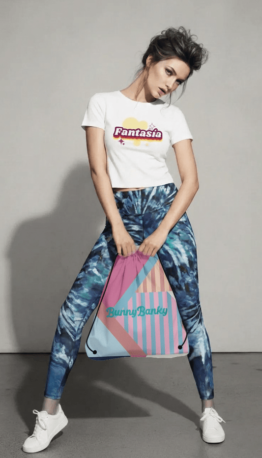 The model is standing in a wide stance, wearing a white "Fantasia" branded T-shirt and blue and green patterned "Athens Sculpt" leggings. She is holding a colorful "BunnyBanky" drawstring bag.