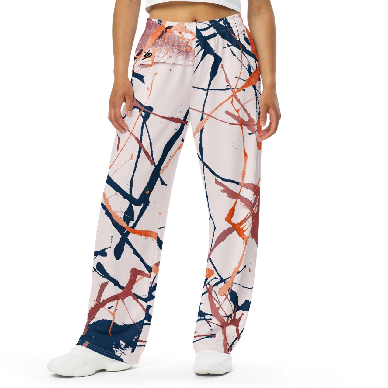 A woman modeling the angel flow wide-leg pants with an abstract paint splatter pattern in navy, orange, and white, paired with a white crop top and white sneakers against a plain white background.