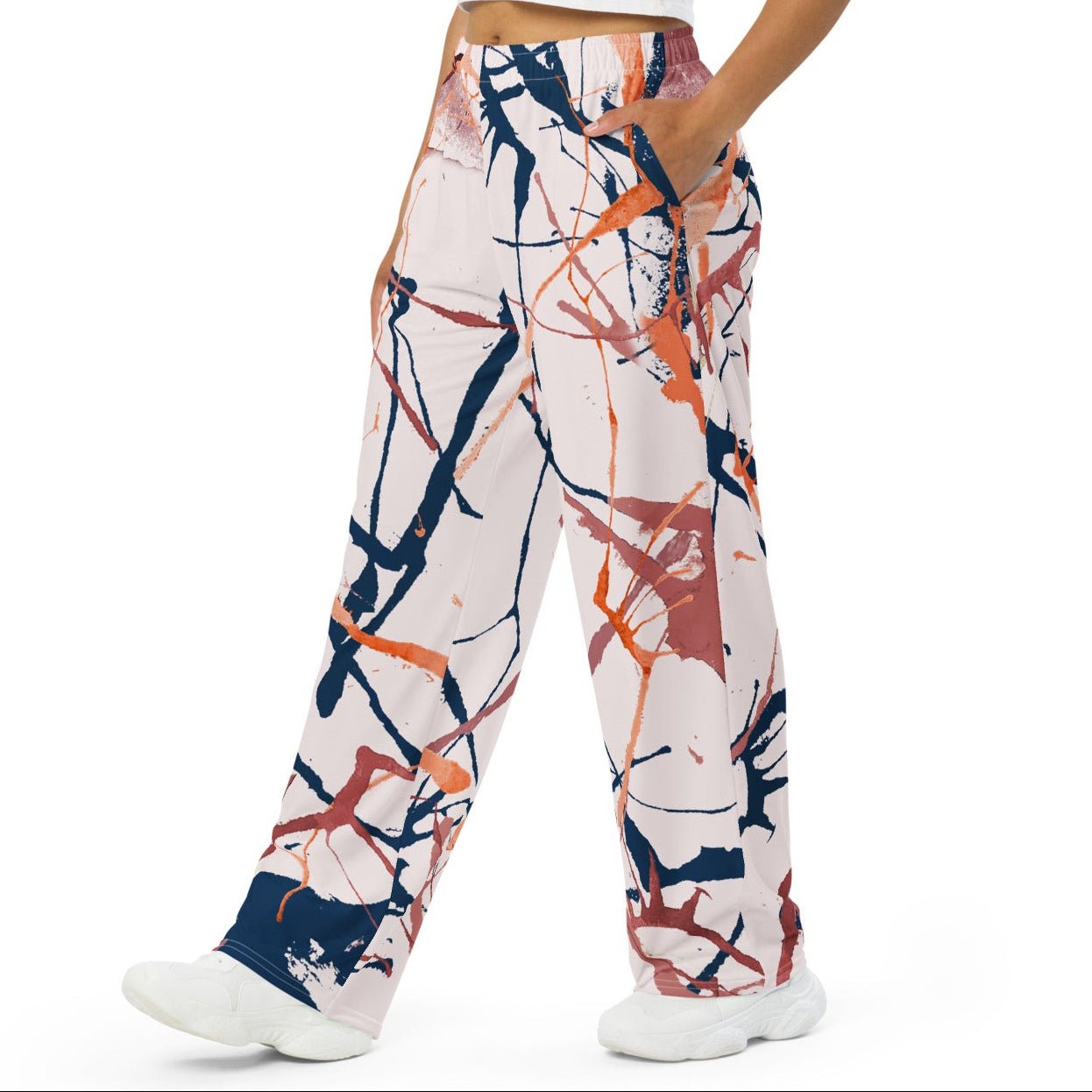 A woman modeling a pair of wide-leg pants with an abstract paint splatter pattern in navy, orange, and brown on a white base, paired with a white crop top and white sneakers against a solid white background.