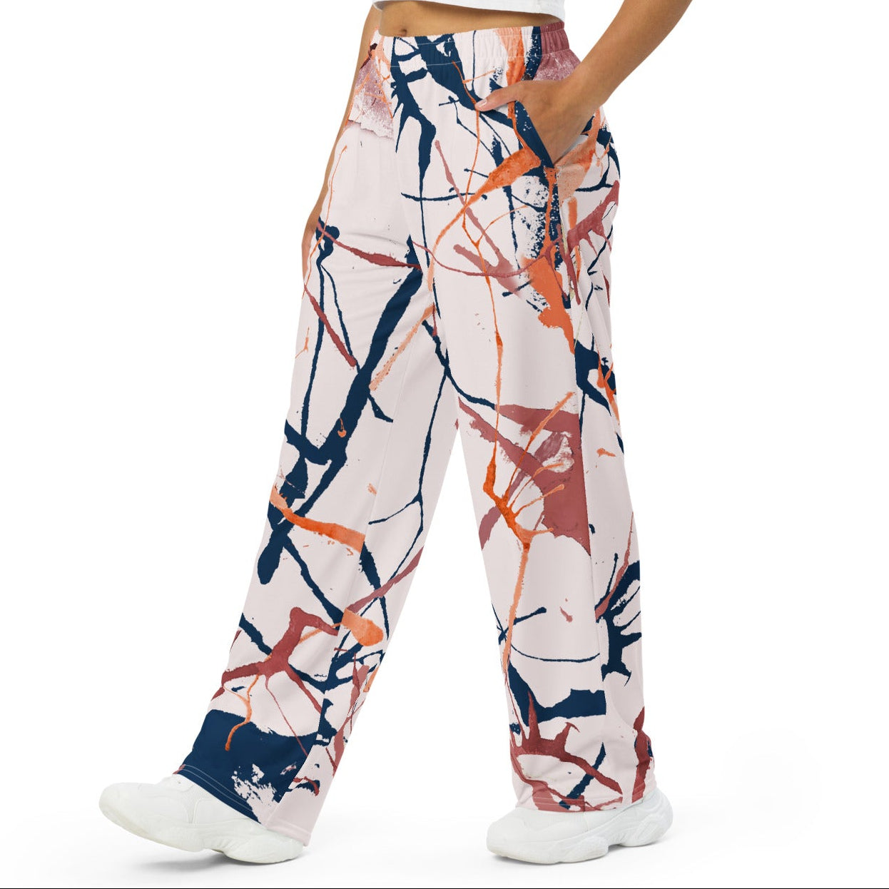 A woman modeling a pair of wide-leg pants with an abstract paint splatter pattern in navy, orange, and brown on a white base, paired with a white crop top and white sneakers against a solid white background.