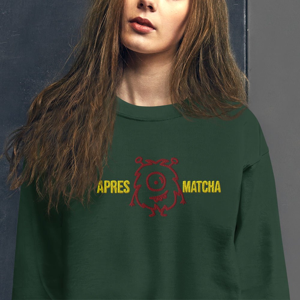 A close-up of a woman wearing a dark green crewneck sweatshirt with a red cartoon monster graphic and yellow text reading 'APRES MATCHA' across the chest, set against a dark gray background.