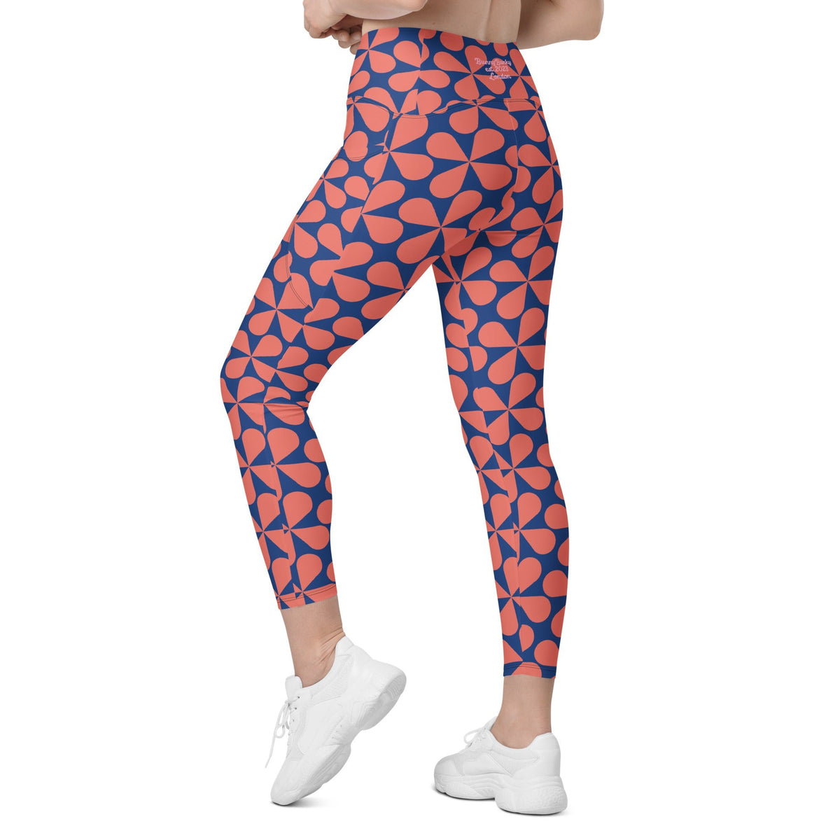Model wearing the  Bakerloo Sculpt Leggings in geometric pattern in blue with coral background.