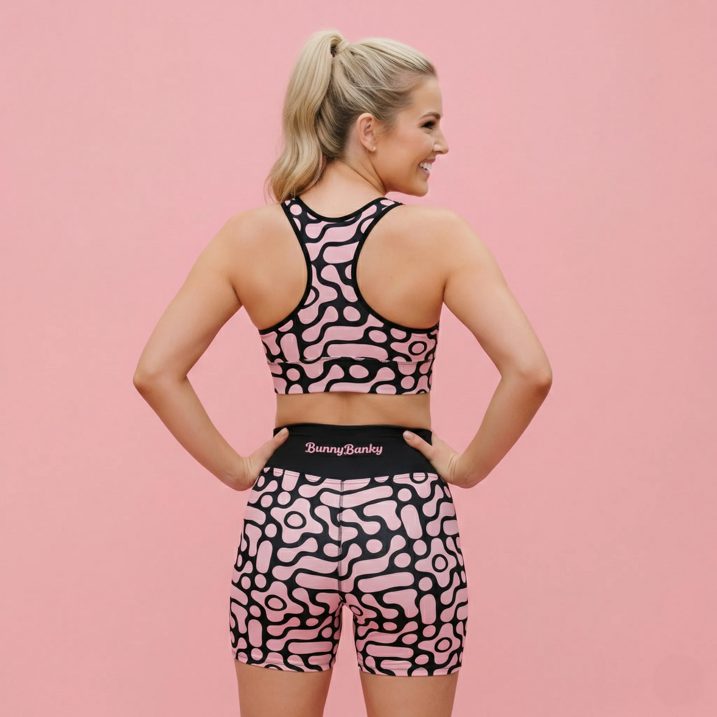 A rear-view studio shot of the BunnyBanky AI proprietary model showcasing the Berlin Performance Bra and matching performance shorts. The set features a unique, custom-designed pattern of geometric black floral shapes set against a vibrant soft pink background. The model is posed with hands on her hips, looking over her shoulder with a smile to highlight the athletic fit and racerback detail of the performance wear.