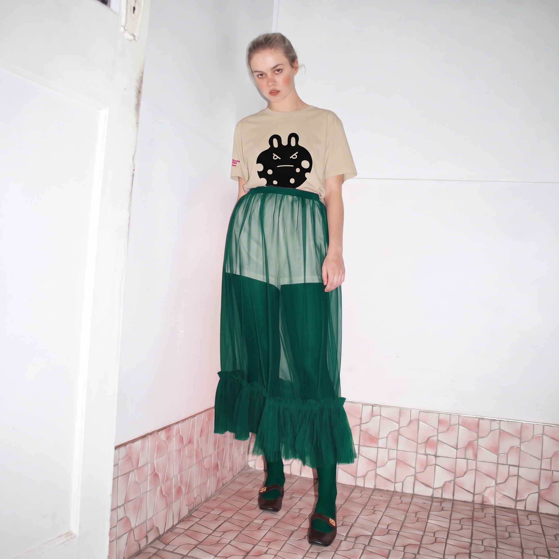 A model stands in a tiled room corner wearing the bunny monster  tee  a desert dust graphic t-shirt with 'bunny monster' printed on the chest, a sheer emerald green mesh skirt over emerald green leggings and black shoes.