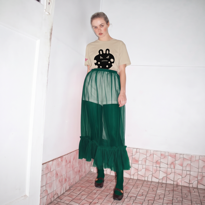 A model stands in a tiled room corner wearing the bunny monster  tee  a desert dust graphic t-shirt with 'bunny monster' printed on the chest, a sheer emerald green mesh skirt over emerald green leggings and black shoes.