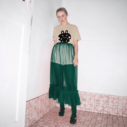 A model stands in a tiled room corner wearing the bunny monster  tee  a desert dust graphic t-shirt with 'bunny monster' printed on the chest, a sheer emerald green mesh skirt over emerald green leggings and black shoes.
