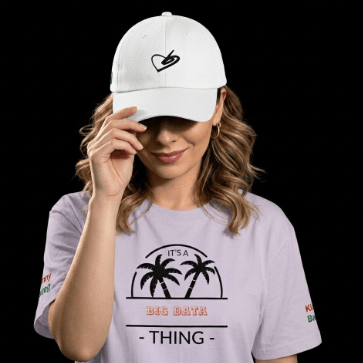 A woman wearing a white baseball cap with a black heart logo and a light lavender graphic T-shirt with palm tree artwork and the text 'IT'S A BEG DATA - THING -' against a solid black background.