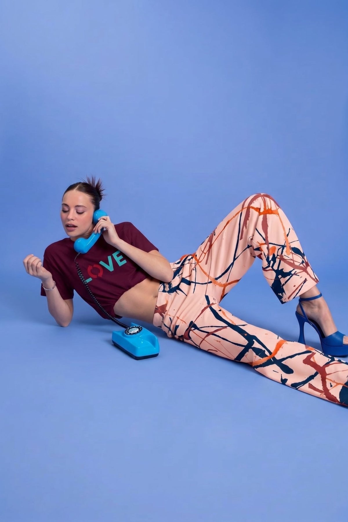 A woman reclines on a solid blue studio floor, holding a bright blue rotary telephone. She is wearing the Love recycled tee, a dark burgundy crop top with colorful 'LOVE' lettering and The angel flow joggers, a pair of wide-leg pants featuring an abstract paint splatter pattern in orange, navy blue, and terracotta on a cream background, shown flat against a white background.