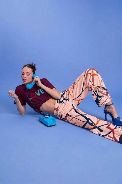 A woman reclines on a solid blue studio floor, holding a bright blue rotary telephone. She is wearing the Love recycled tee, a dark burgundy crop top with colorful 'LOVE' lettering and The angel flow joggers, a pair of wide-leg pants featuring an abstract paint splatter pattern in orange, navy blue, and terracotta on a cream background, shown flat against a white background.