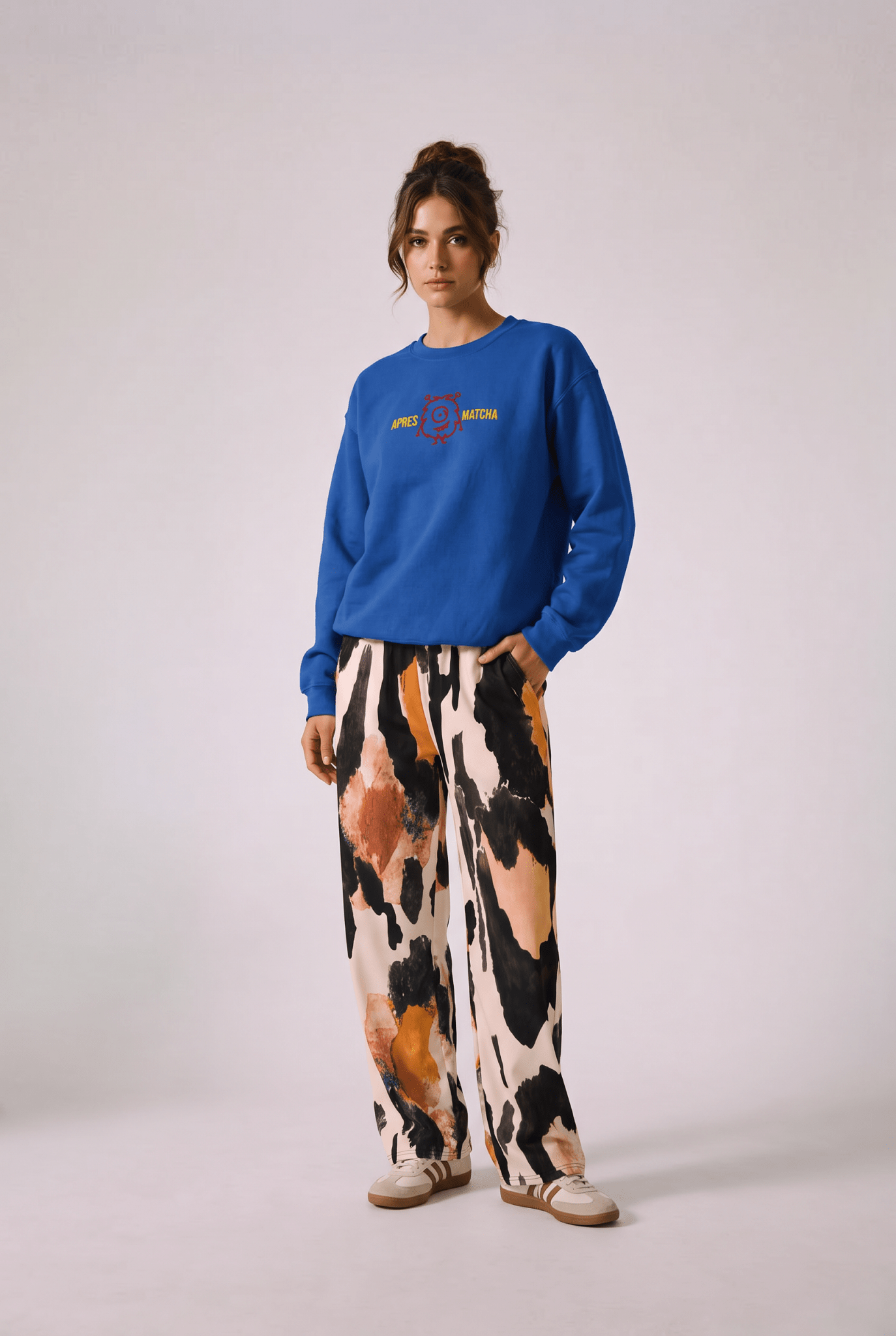 A model wearing  the apres matcha sweatshirt in  royal blue with a colorful graphic on the chest, and the amazonia flow joggers, a wide-leg animal patterned pants and sneakers against a plain studio background.