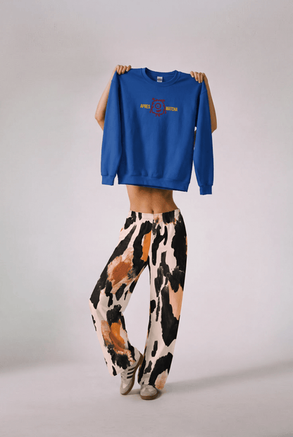 A model holds up the apres matcha sweatshirt in  royal blue with a colorful graphic on the chest, while wearing the amazonia flow joggers, a wide-leg animal patterned pants and sneakers against a plain studio background.