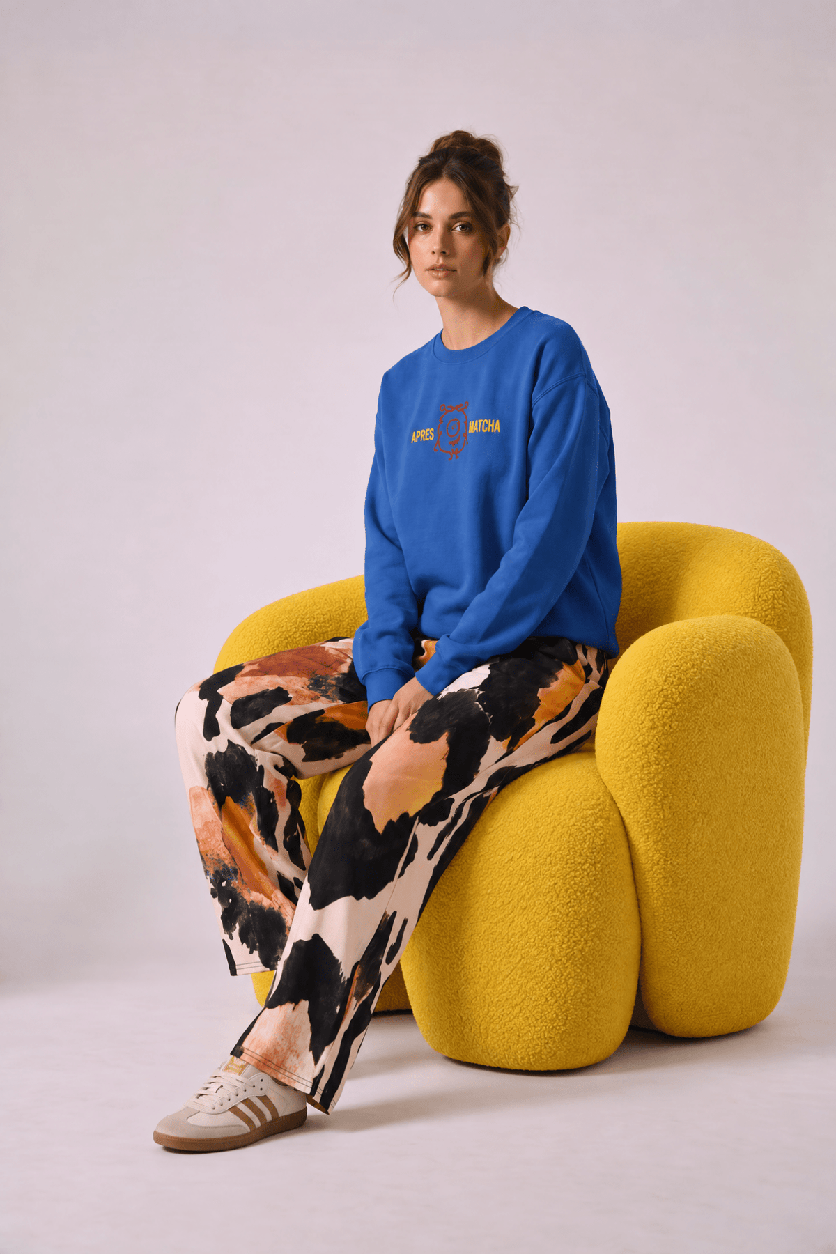 A model sitting on a sculpted yellow chair wearing  the apres matcha sweatshirt in  royal blue with a colorful graphic on the chest, and the amazonia flow joggers, a wide-leg animal patterned pants and sneakers against a plain studio background.