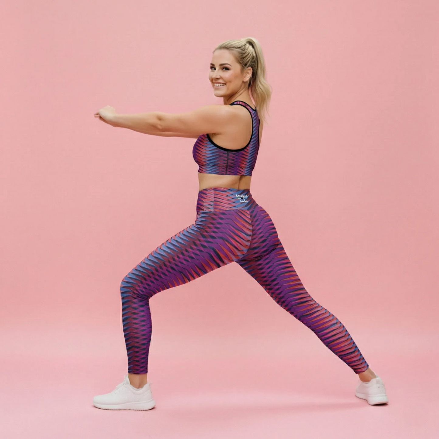 A BunnyBankY AI proprietary fitness model with a blonde ponytail is captured in a dynamic side-profile lunge, looking back with a smile against a soft pink background. She is wearing the Moorgate Performance Bra and Moorgate Sculpt Leggings, which feature a vibrant, repeating geometric arrow print in shades of pink, orange, and blue. The high-waisted leggings are designed for active performance with mild compression and include functional dual side pockets.