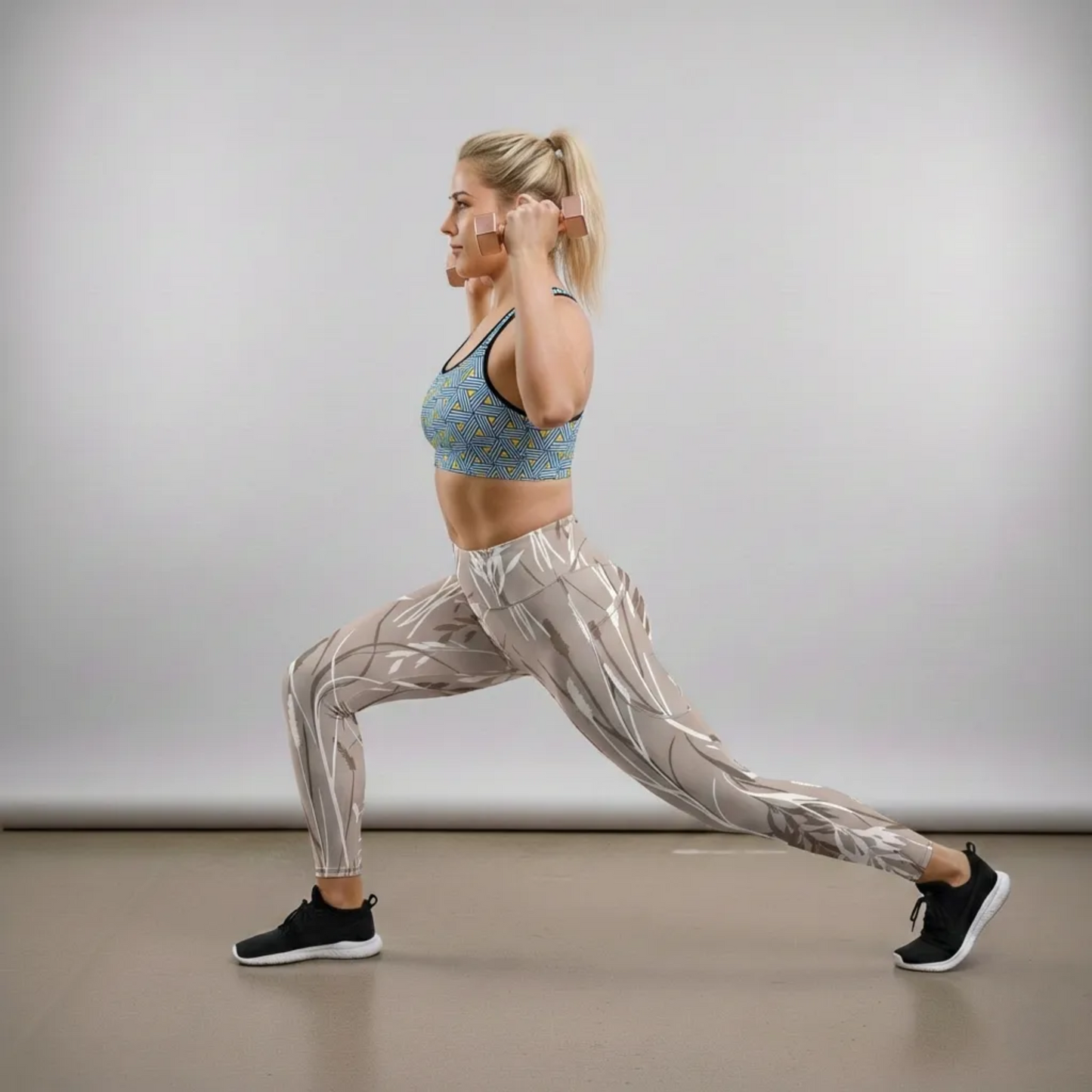 A blonde BunnyBankY AI proprietary fitness model performs a deep lunge in a studio setting, holding rose gold dumbbells. She is wearing the Geneva Performance Bra, which features a blue and yellow geometric triangular pattern, paired with the Osaka Sculpt Leggings, which have a neutral taupe base with an abstract white and grey botanical line art design.