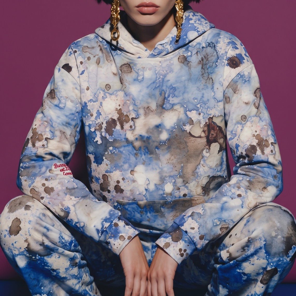 A model wearing the Amalfi Resort Hoodie and matching flow joggers in a matching blue and white abstract print hoodie and sweatpants set, posing in a crouched position against a solid purple background.