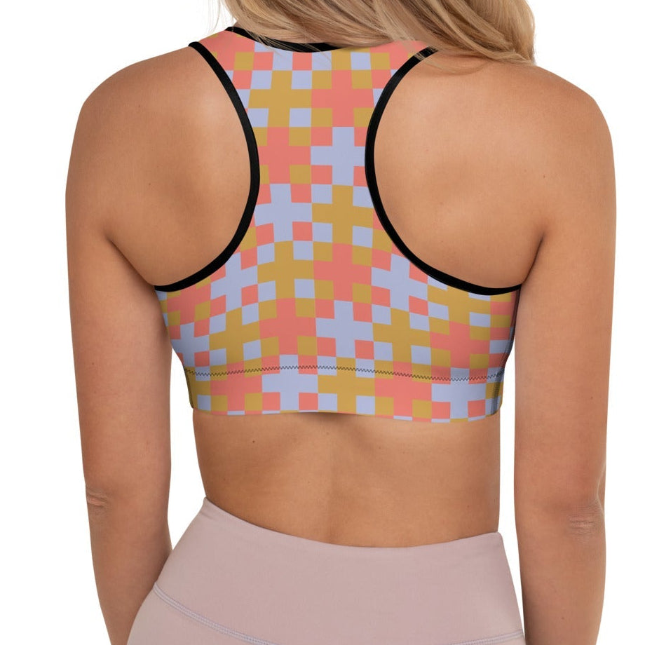 A  model wearing the Pixel Glam Performance Bra, highlighting the geometric scoop neckline, wide under-bust band, and vibrant terracotta, gold, and blue pixelated grid pattern.