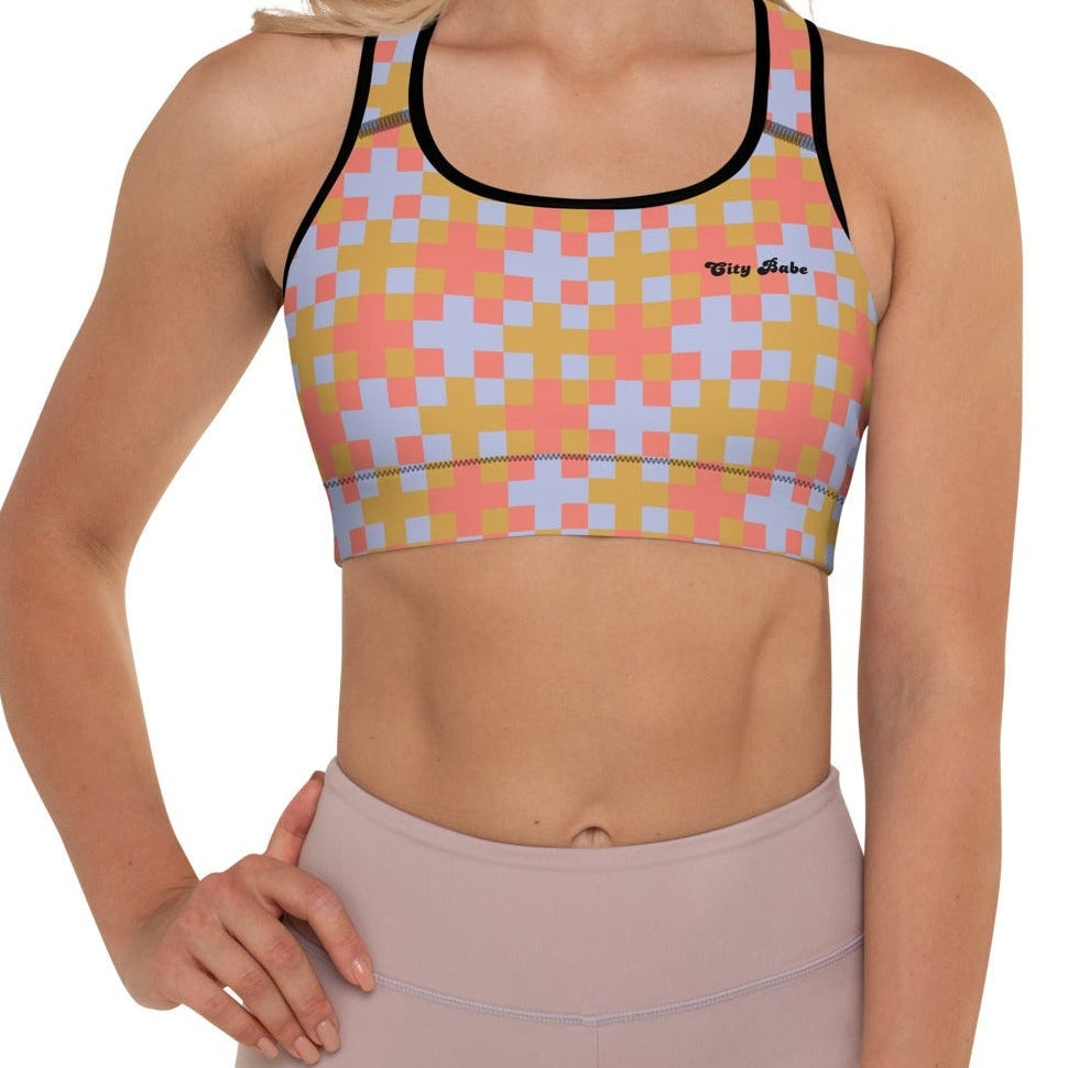 A  model wearing the Pixel Glam Performance Bra, highlighting the geometric scoop neckline, wide under-bust band, and vibrant terracotta, gold, and blue pixelated grid pattern.