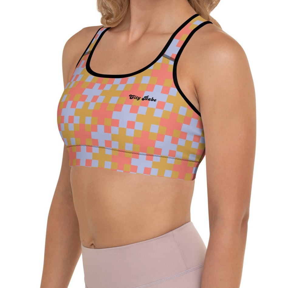 A  model wearing the Pixel Glam Performance Bra, highlighting the geometric scoop neckline, wide under-bust band, and vibrant terracotta, gold, and blue pixelated grid pattern.