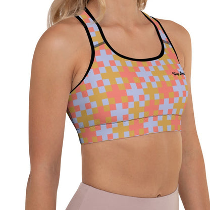 A  model wearing the Pixel Glam Performance Bra, highlighting the geometric scoop neckline, wide under-bust band, and vibrant terracotta, gold, and blue pixelated grid pattern.
