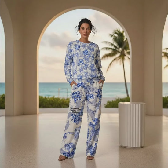 A woman stands in a modern, arched terrace overlooking a tropical beach at sunset, wearing the Saint Germain Glam Co-ord set. The outfit features a white jumper and matching wide-leg joggers adorned with an intricate, vintage-inspired blue floral and toile-like pattern.