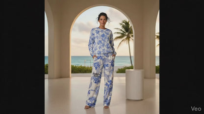 A woman stands in a modern, arched terrace overlooking a tropical beach at sunset, wearing the Saint Germain Glam Co-ord set. The outfit features a white jumper and matching wide-leg joggers adorned with an intricate, vintage-inspired blue floral and toile-like pattern.
