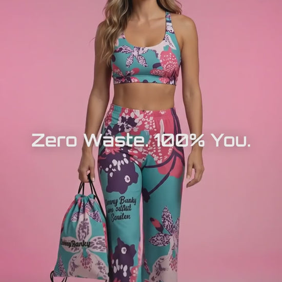 Vertical video of model Athena wearing the BunnyBanky Signature Frequency Glam Set, featuring a high-performance bra, wide-leg Chelsea flow joggers, and a matching drawstring backpack in a bold, vibrant teal, pink, and purple floral pattern against a soft studio pink background.