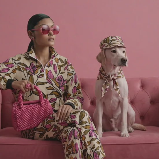A model wearing the terra flow joggers and shell, a beige floral print tracksuit, pink sunglasses, and a pink beaded necklace sits on a pink velvet sofa next to a  dog in a matching outfit.