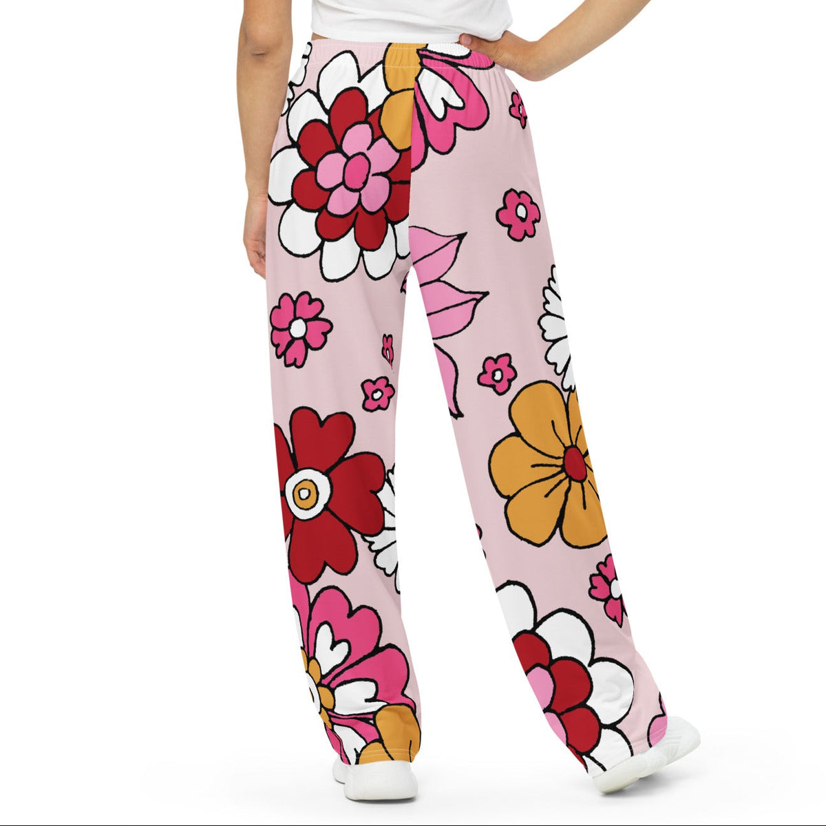 A model wearing the primrose hill flow joggers. pink wide-leg pants with a retro floral print, styled with a white crop top and white sneakers against a plain white background.