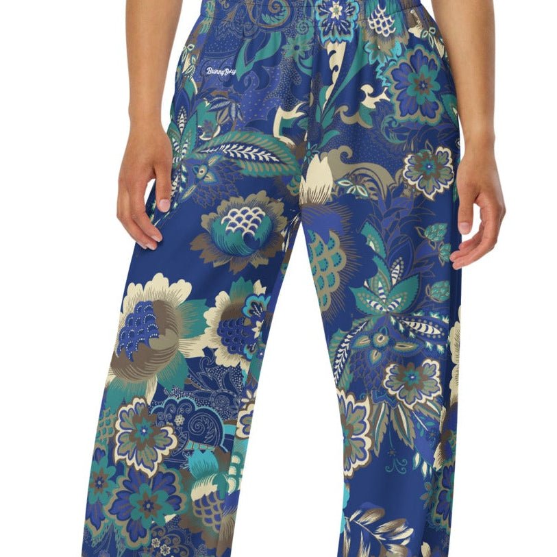 A woman wearing the rio flow joggers, wide-leg cut with a detailed botanical pattern in blue, green, and gold, paired with a white t-shirt and white sneakers, set against a plain white background.