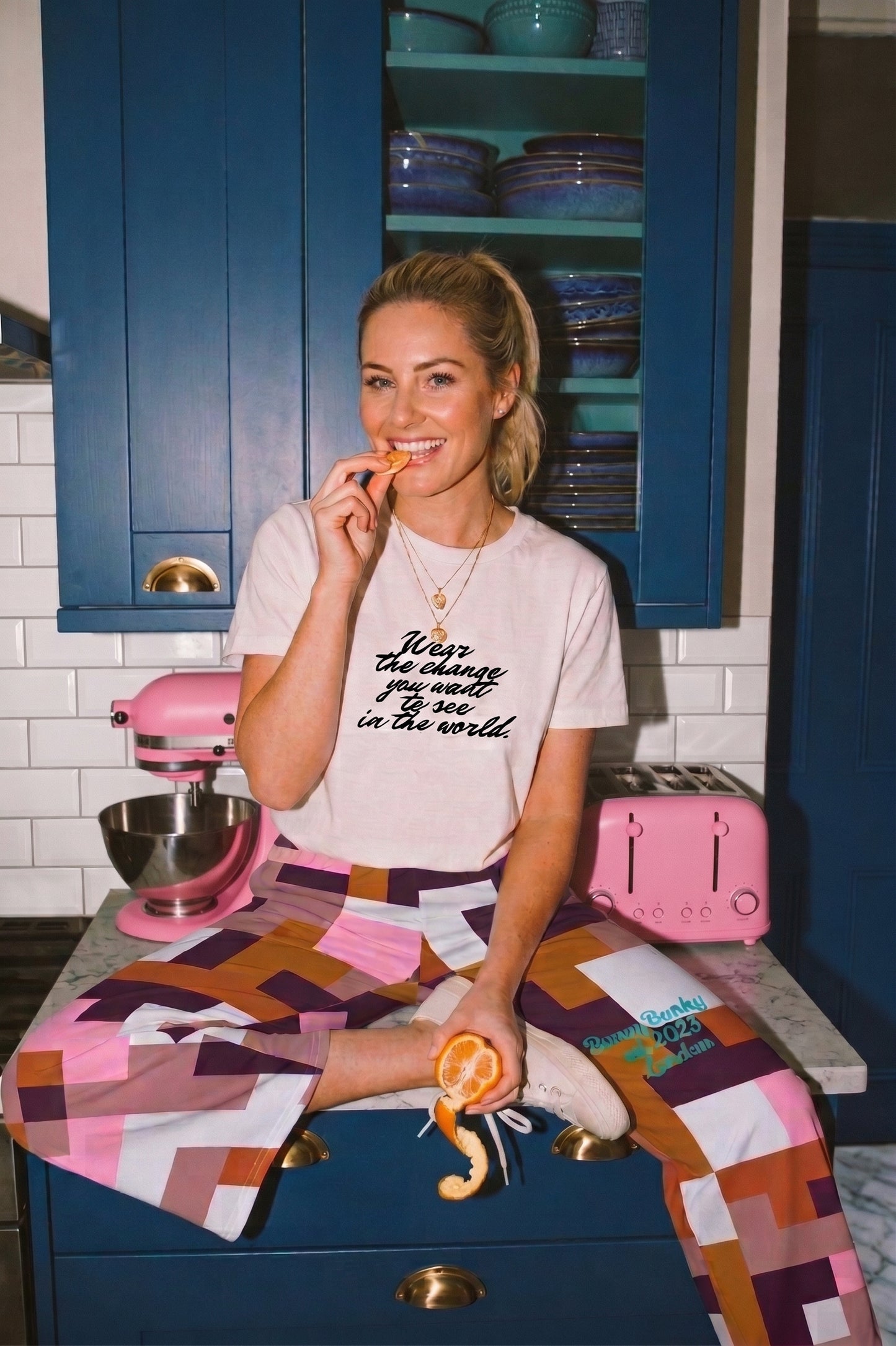 BunnyBanky AI proprietary model sitting cross-legged on a navy blue kitchen counter holding a peeled tangerine. She wears the BunnyBanky white organic cotton graphic tee with black script reading "Wear the change you want to see in the world" and the Marais Flow Joggers featuring a pink, brown, and plum geometric block pattern. Background shows white subway tiles, a pink retro stand mixer, and a pink toaster.