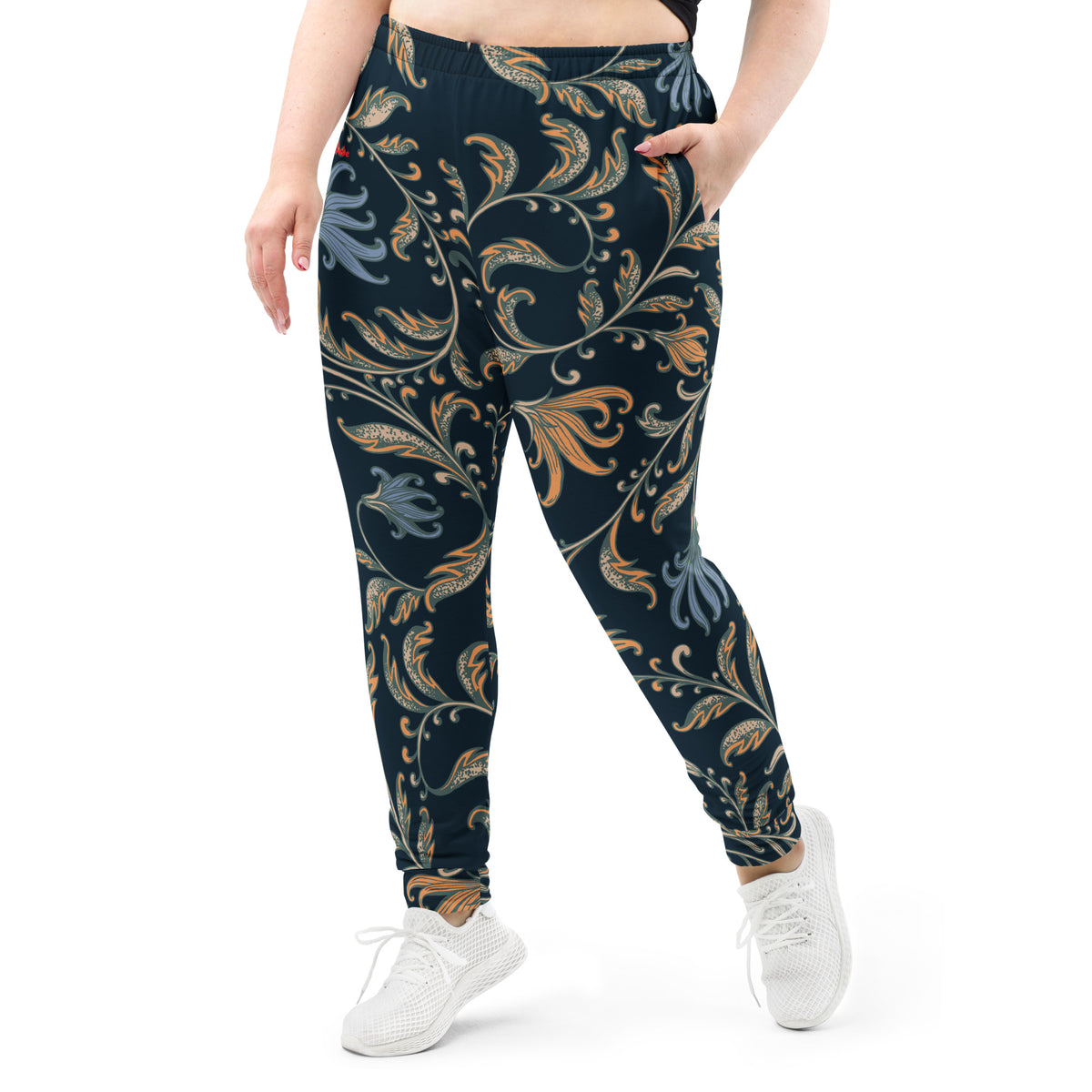 A model wearing the Venice slim fit high-waisted athletic joggers with a dark navy blue background and an intricate botanical pattern in gold, beige, and light blue. The joggers feature a small red logo on the left hip.