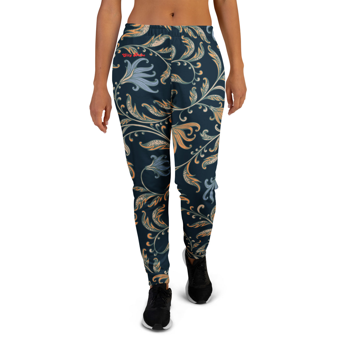 A model wearing the Venice slim fit high-waisted athletic joggers with a dark navy blue background and an intricate botanical pattern in gold, beige, and light blue. The joggers feature a small red logo on the left hip.